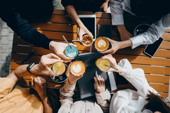 Top-down view of people clinking various iced and hot coffee drinks together over a wooden outdoor table.
