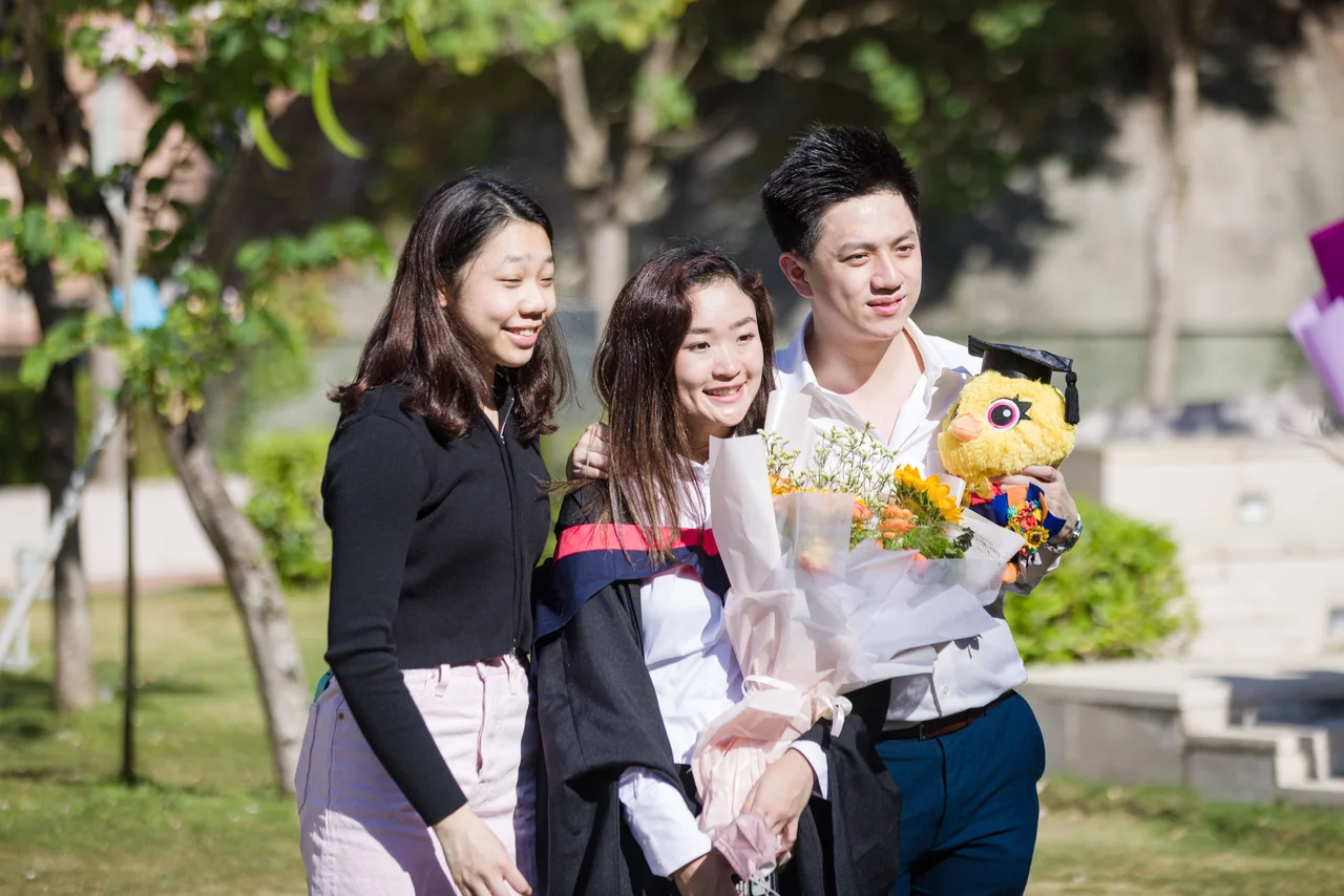 A female graduate holding flowers poses outdoors with two friends, one of whom holds a plush toy wearing a graduation cap.