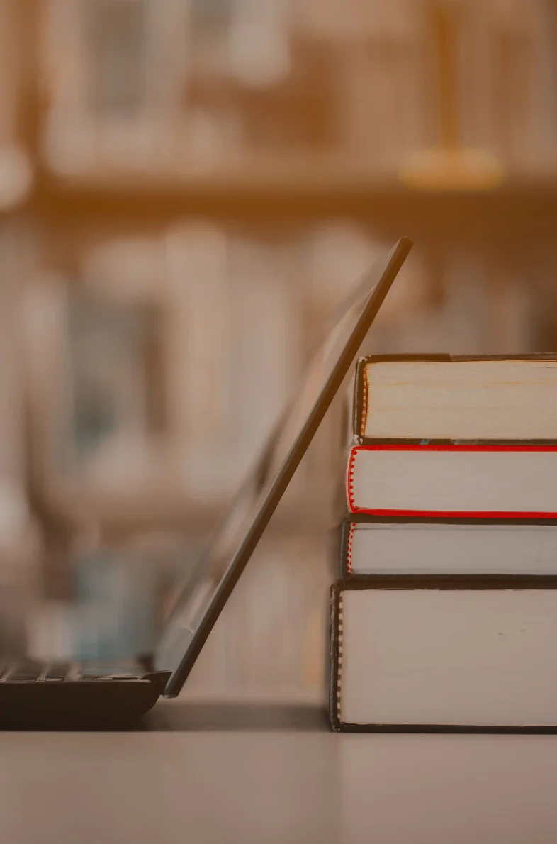 Side view of an open laptop screen leaning against a stack of four thick books in a blurred library setting.