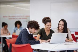 Three students working together around a table with a laptop, while other students study in the background classroom.