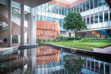 A modern campus courtyard featuring a reflecting pool with clear reflections of a brick wall displaying the HKU coat of arms and text, surrounded by glass-facade buildings, trees, and a green lawn.