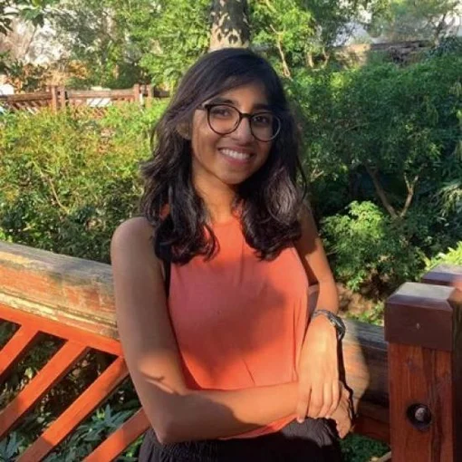 A smiling young woman with glasses and wavy dark hair wearing an orange sleeveless top, leaning against a wooden railing outdoors surrounded by green plants.