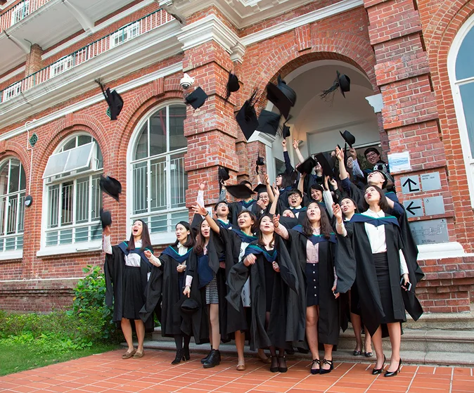 Graduates in academic gowns throwing mortarboards into the air outside HKU Main Building's red-brick arched doorway.