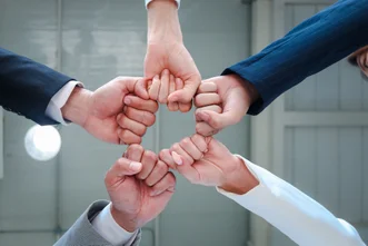 Direct overhead view of five people in business suits joining their fists together in a star-shaped circle.