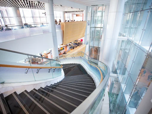 Curved staircase inside modern multi-story university building with glass walls and study areas.
