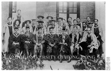 Historical black-and-white group portrait of staff and graduates wearing academic gowns seated and standing outdoors in front of a building.
