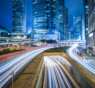 Long exposure photograph of light trails from traffic on multi-lane city roads leading into an underpass amidst illuminated skyscrapers at night.