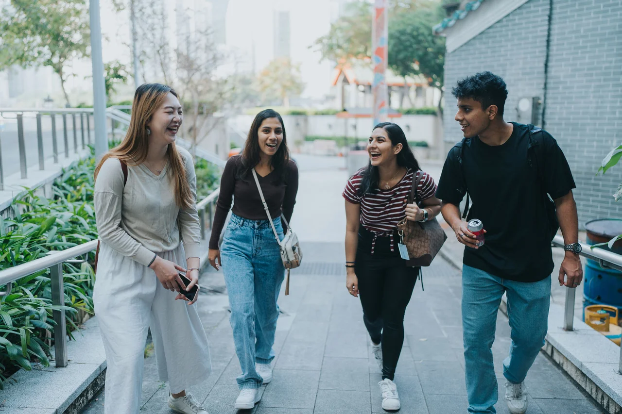 Four young adults laughing and chatting together while walking along a paved outdoor walkway.