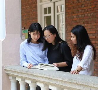Three women reading a book together along an outdoor corridor with white balustrades and red-brick walls.