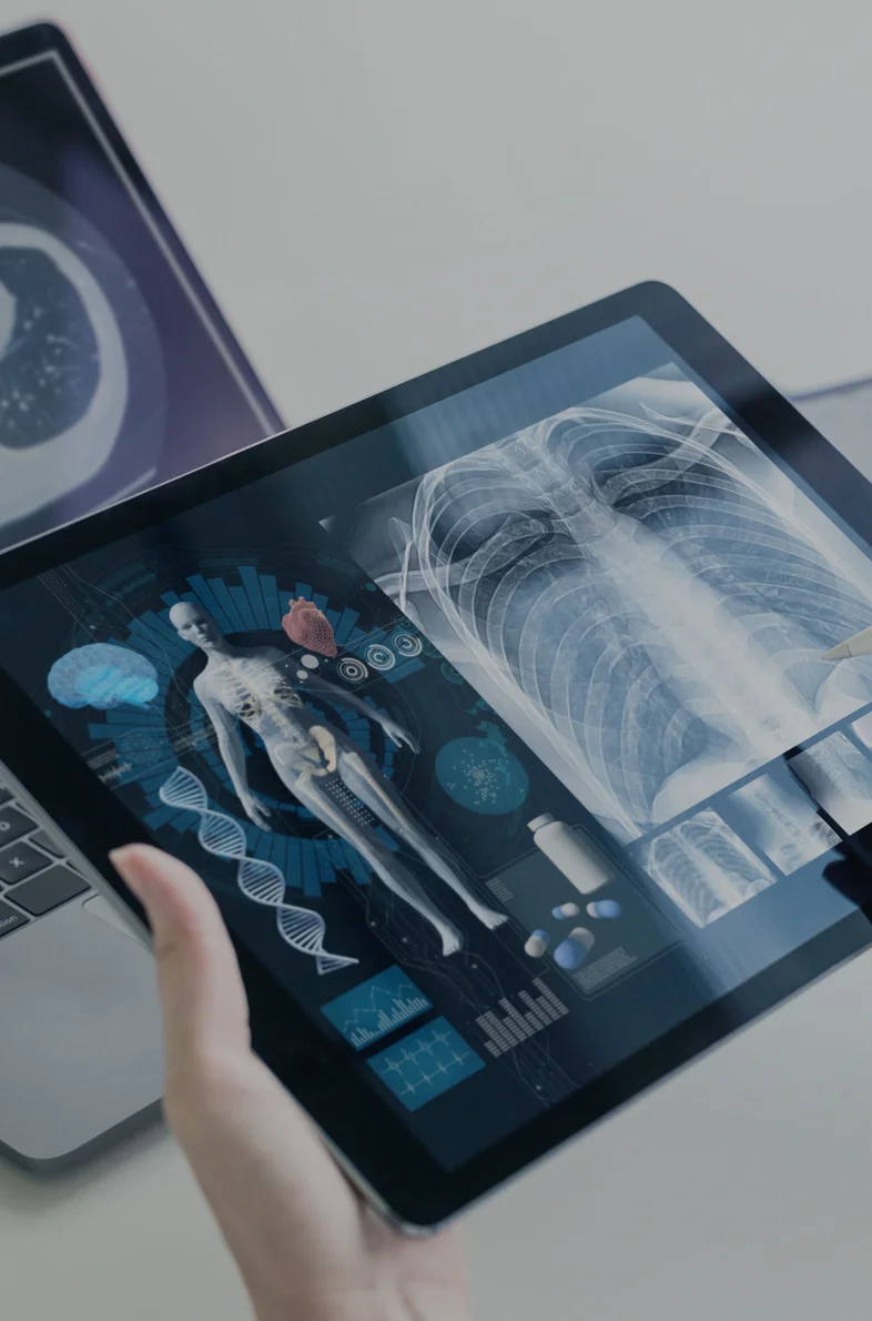 A person holding a stylus points at chest X-rays on a tablet, with a laptop displaying a CT scan nearby.