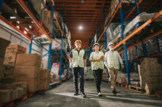 Three warehouse workers in hard hats and safety vests walking down an aisle lined with stacked shelving units.