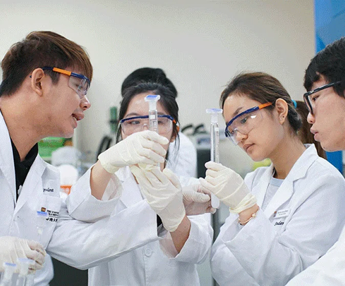 HKU students wearing lab coats, safety goggles, and gloves inspect test tubes together in a laboratory setting.