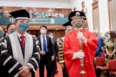 HKU President Xiang Zhang wearing academic regalia and a face mask walks alongside a mace-bearer holding the silver-gilt ceremonial mace during a indoor graduation procession.