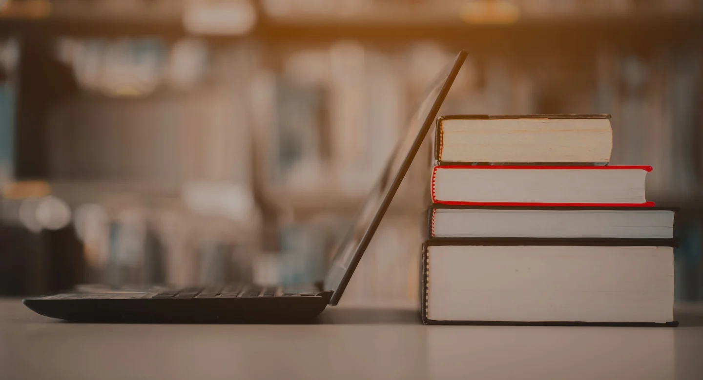 Side view of an open laptop screen leaning against a stack of four thick books in a blurred library setting.