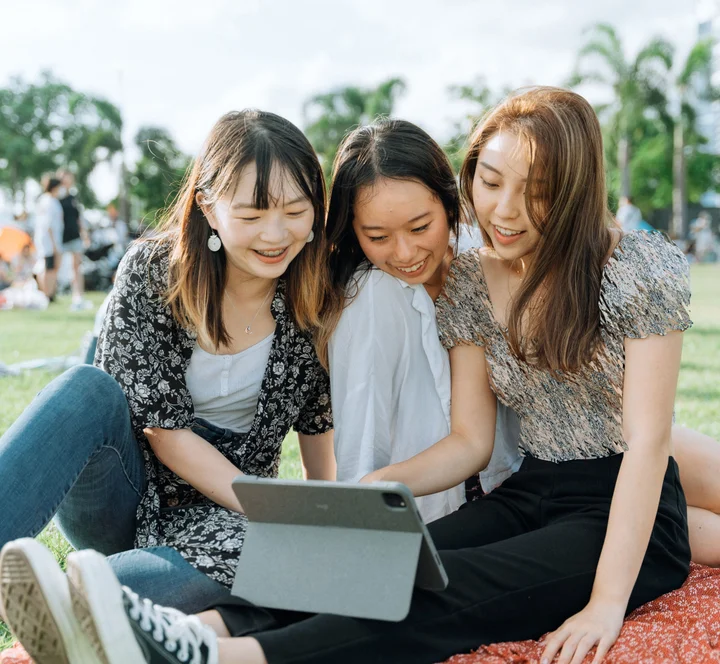Three smiling young women sitting on a lawn outdoors and looking at a tablet together.