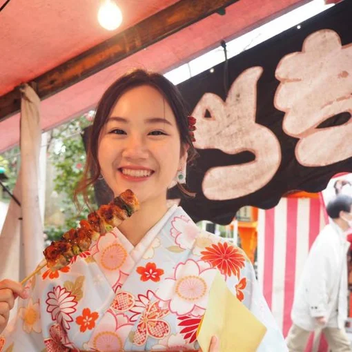 A smiling young woman in a floral kimono holding a skewer of grilled food at an outdoor market stall.