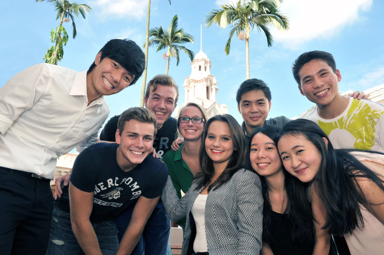 A group of smiling students pose closely together outdoors, with palm trees and HKU Main Building's clock tower in the background.