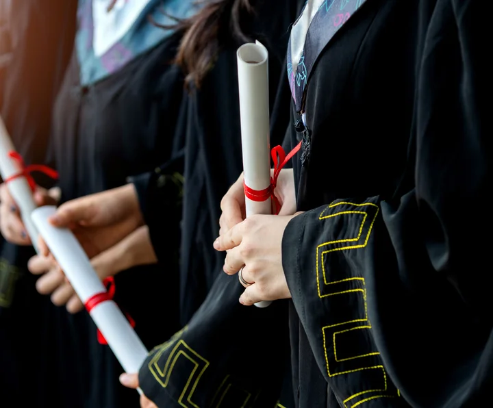 Close-up of graduates in black academic gowns holding rolled diplomas tied with red ribbons.