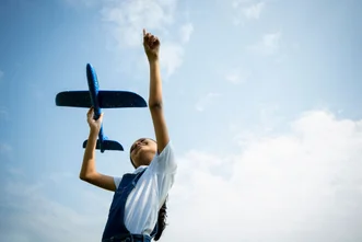 Low-angle shot of a young girl holding a blue toy airplane up toward a cloudy blue sky while pointing upward.