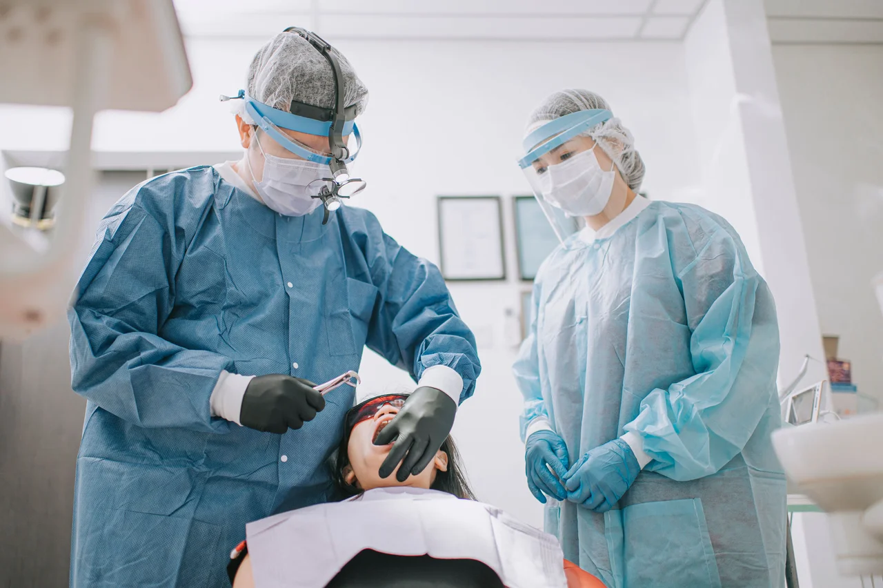 Dentist holding dental pliers treats a patient lying in a chair, assisted by a healthcare worker in protective gear.