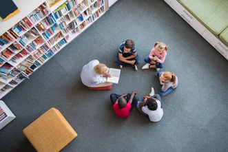 Top-down view of a teacher reading a book to five young children seated in a circle on a carpeted library floor.
