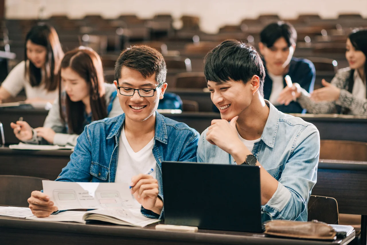 Two male students discussing notes and a laptop screen while seated in a lecture hall.