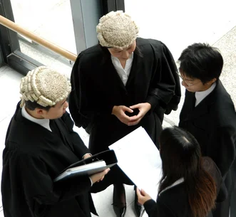 High-angle view of four legal professionals in black robes—two wearing traditional wigs—standing in a circle discussing documents.