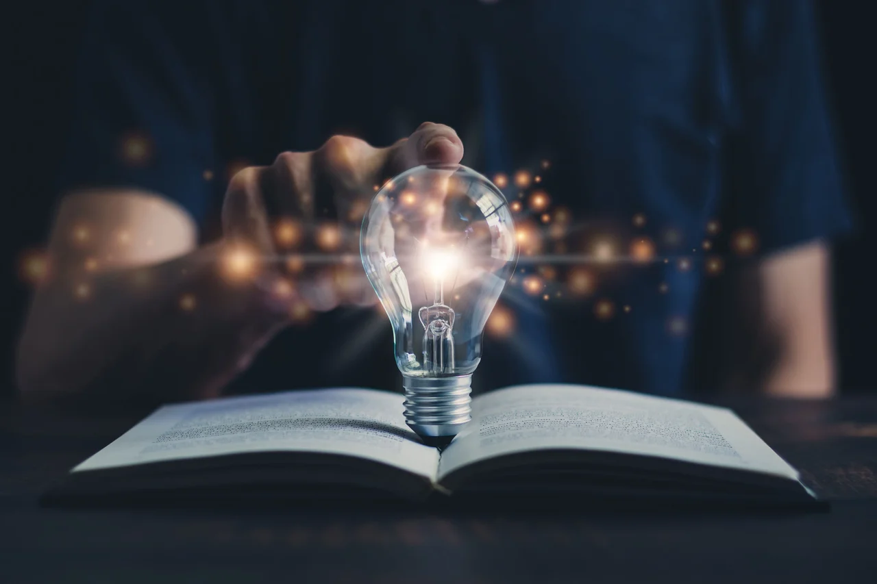 Hand reaching toward a glowing light bulb resting on an open book. 