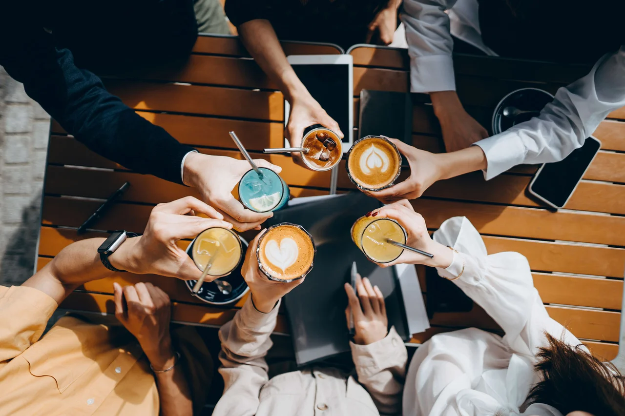 Top-down view of people clinking various iced and hot coffee drinks together over a wooden outdoor table.