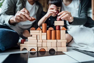 A child building a structure out of wooden blocks with help from two adults sitting on the floor indoors.