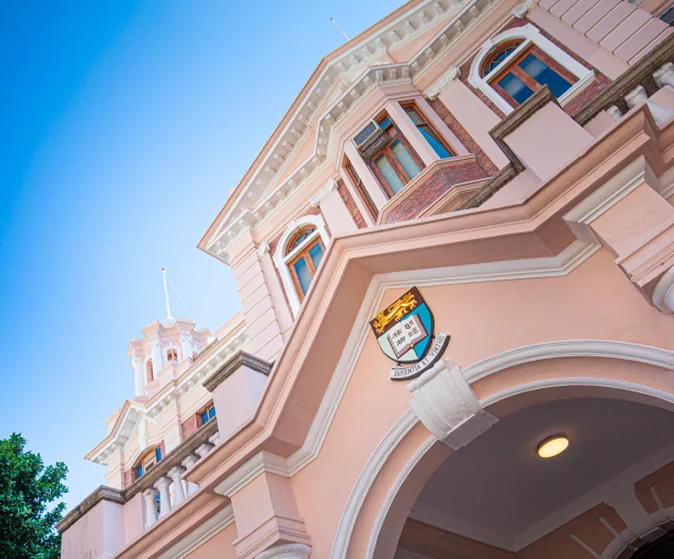 Low-angle view of HKU Main Building facade with the university coat of arms mounted above an arched entrance under a clear blue sky.