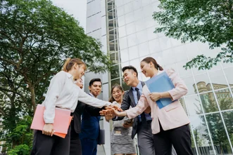 Low-angle view of six young professionals holding folders and stacking their hands together outdoors in front of a glass building.