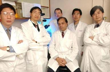 A group of six researchers wearing white laboratory coats posed together inside a laboratory setting with equipment in the background.