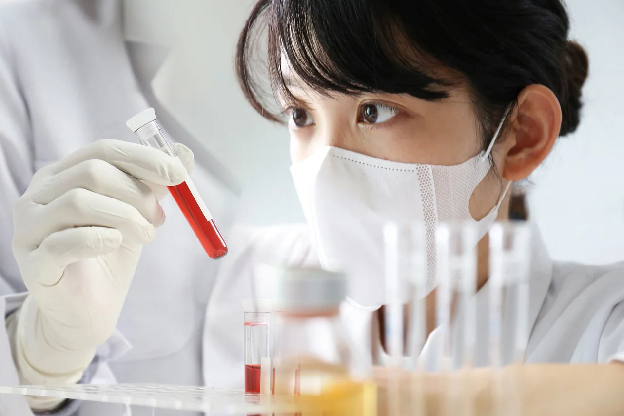A researcher wearing a face mask and gloves closely examines a test tube filled with a red liquid sample in a laboratory.