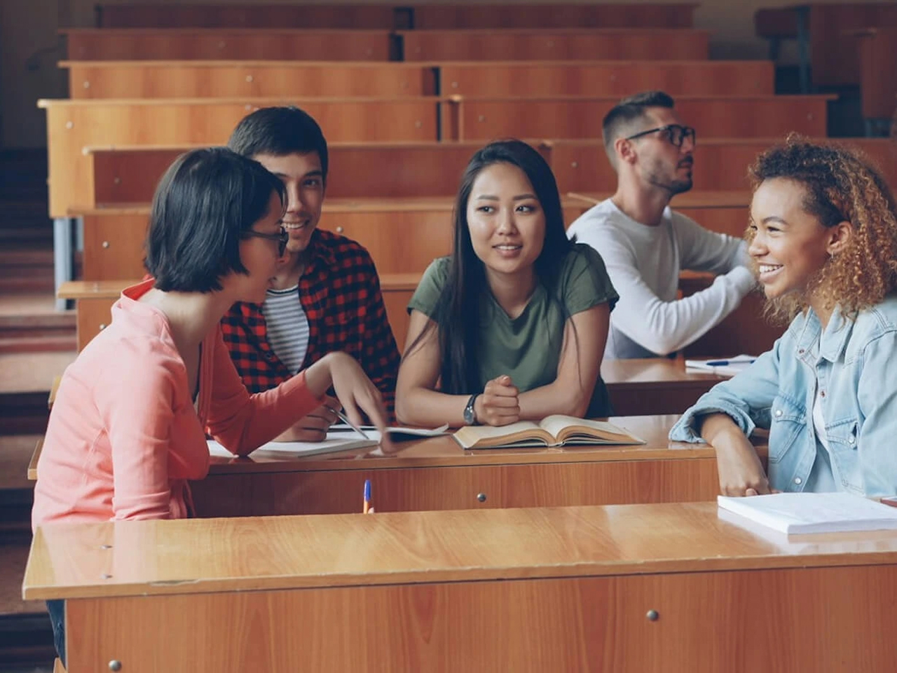 Students sitting at wooden desks in a lecture hall chatting and studying together.